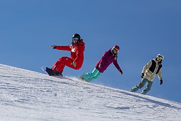 Three snowboarders are riding down a snow covered slope and one of them is wearing a red jacket with the letter h on it