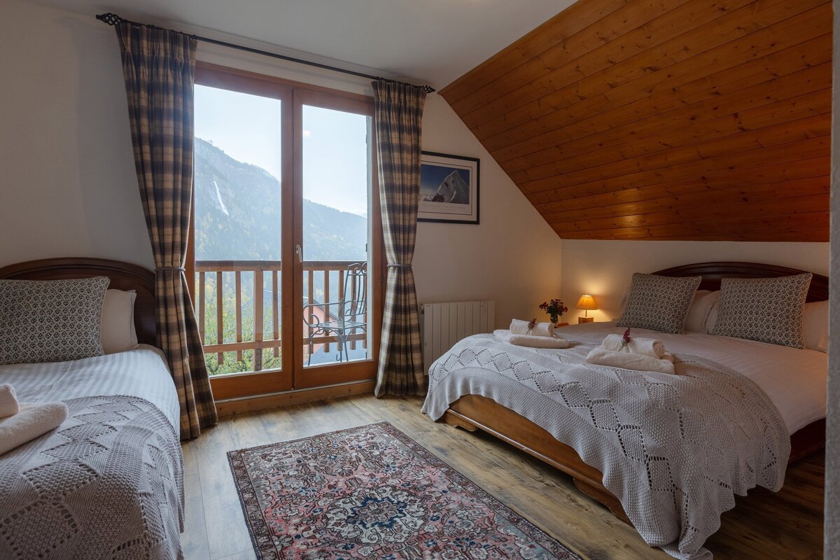 Cozy bedroom with two beds, a patterned rug, and a balcony offering stunning mountain views. Features a warm wooden ceiling and floor.