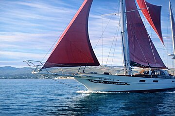 A white sailboat with vibrant red sails glides across calm blue waters, backed by distant mountains and a blue sky.