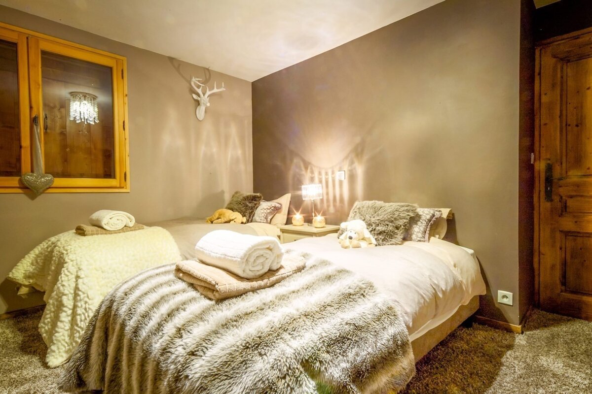 A cozy twin bedroom with faux fur blankets, warm lighting, and a deer head decoration. It has a rustic chalet feel with wooden accents.