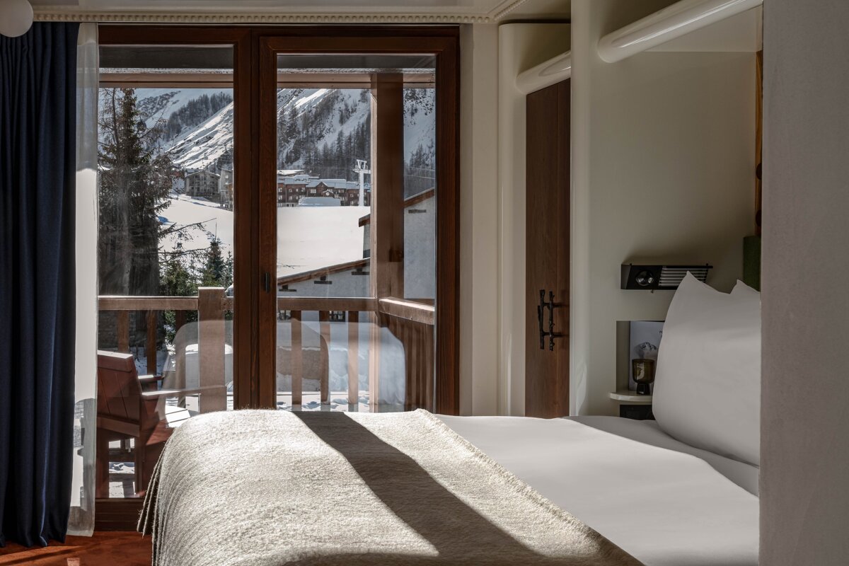 A cozy bedroom with a bed by a balcony, revealing a picturesque snowy mountain village and ski slopes outside.