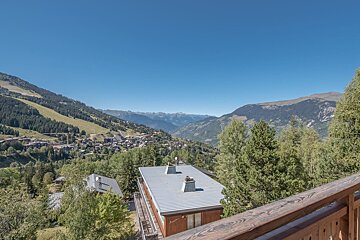 Scenic mountain village vista with lush green trees under a clear blue sky, viewed from a balcony.