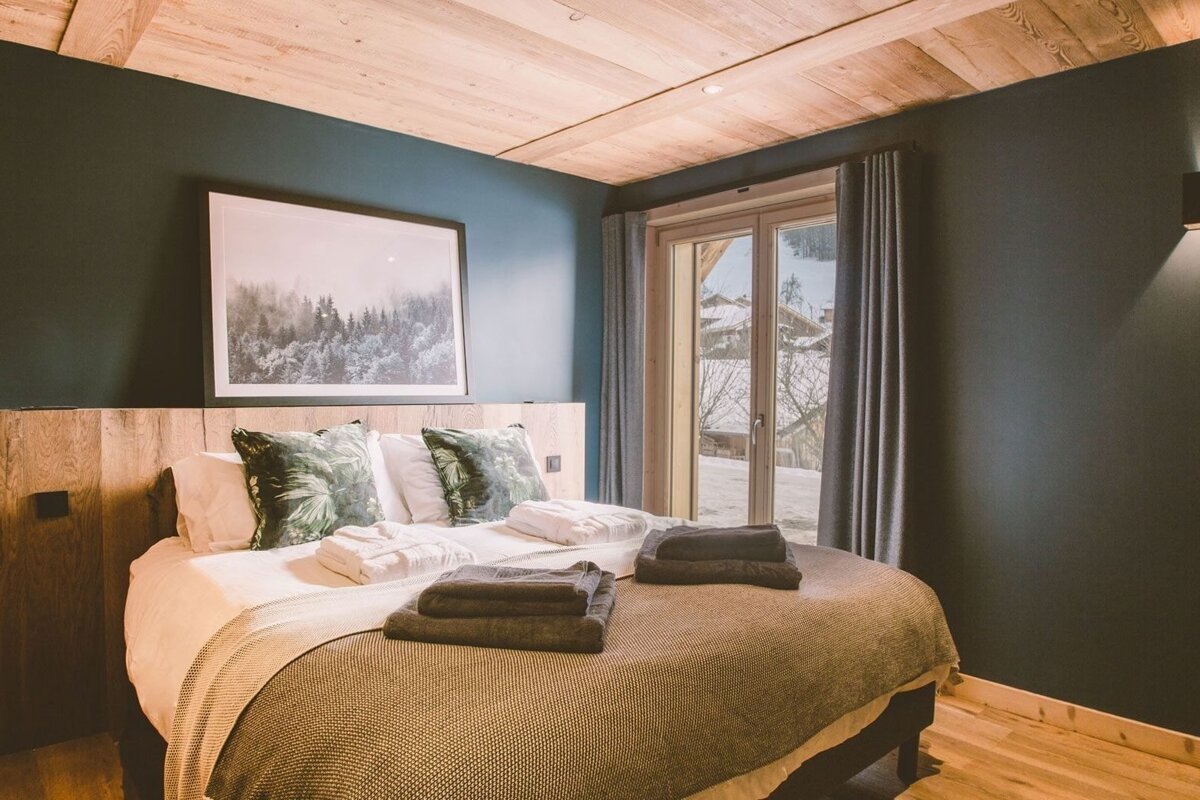 A cozy bedroom with dark teal walls, a wooden ceiling, and a bed with green pillows. A large window offers a snowy winter mountain view.