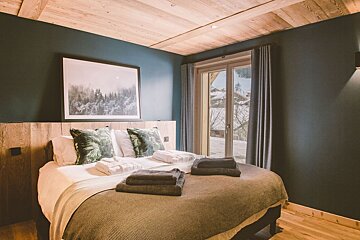 A cozy bedroom with dark teal walls, a wooden ceiling, and a bed with green pillows. A large window offers a snowy winter mountain view.