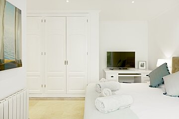 Bright white bedroom with a large built-in wardrobe, a bed with rolled towels and blue pillows, and a TV on a desk.