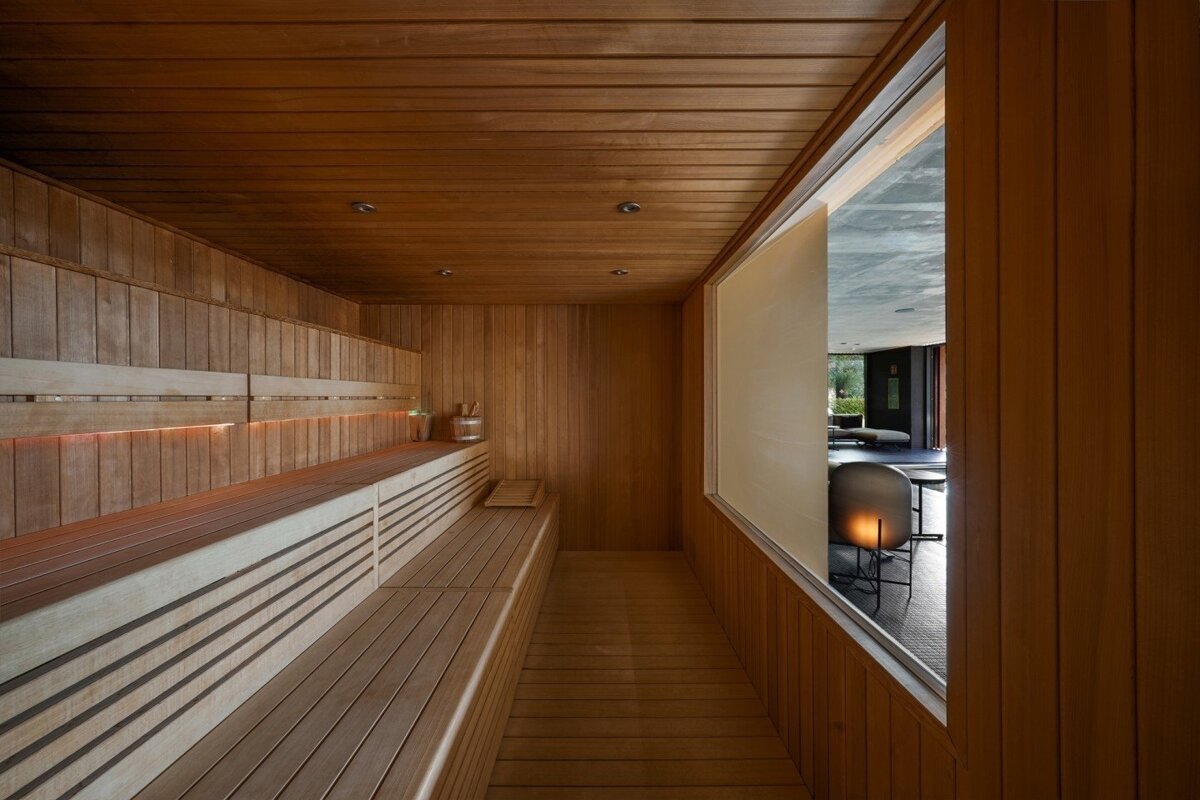 A modern wooden sauna with multi-tiered benches and subtle lighting, offering a view through a large window into a contemporary lounge area.