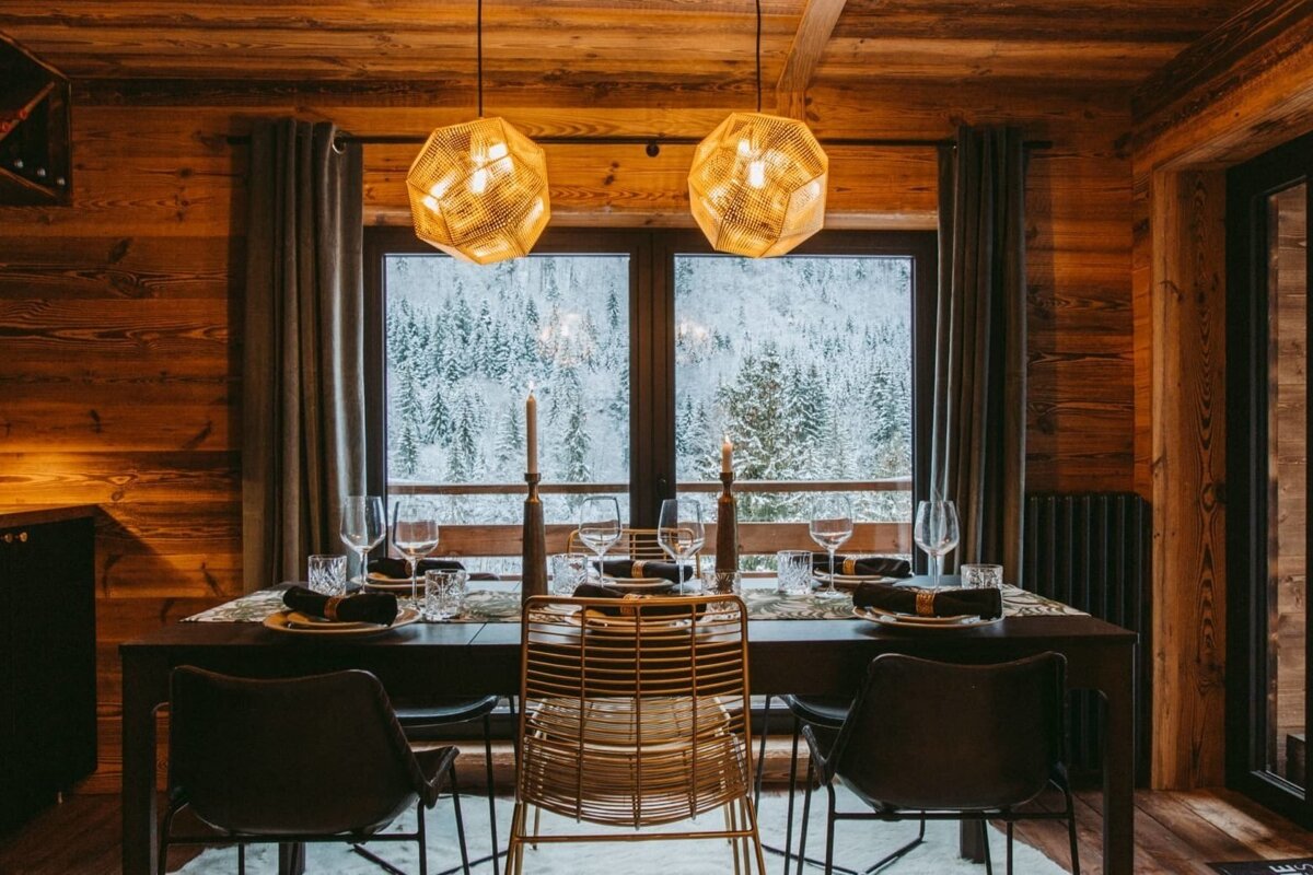 A cozy, rustic wooden dining room features a set table with warm lighting. A large window offers a beautiful view of a snowy forest.