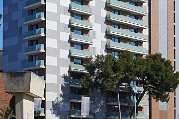 A modern high-rise building with light blue glass balconies and a gray/white geometric facade, partially obscured by green trees in the foreground.