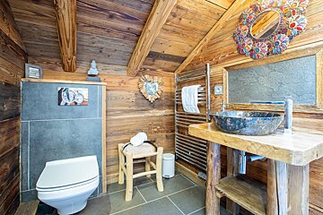 Cozy, rustic wood-paneled bathroom with a unique stone sink, modern toilet, ornate mirror, and wood beam ceiling. A warm chalet feel.