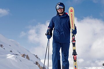 A skier in a blue snowsuit, helmet, and goggles stands on a snowy mountain holding yellow skis and ski poles.