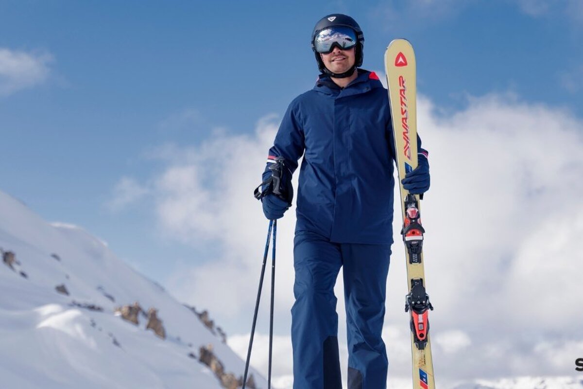 A skier in blue gear stands on a snowy slope holding a ski and poles under a partly cloudy blue sky.