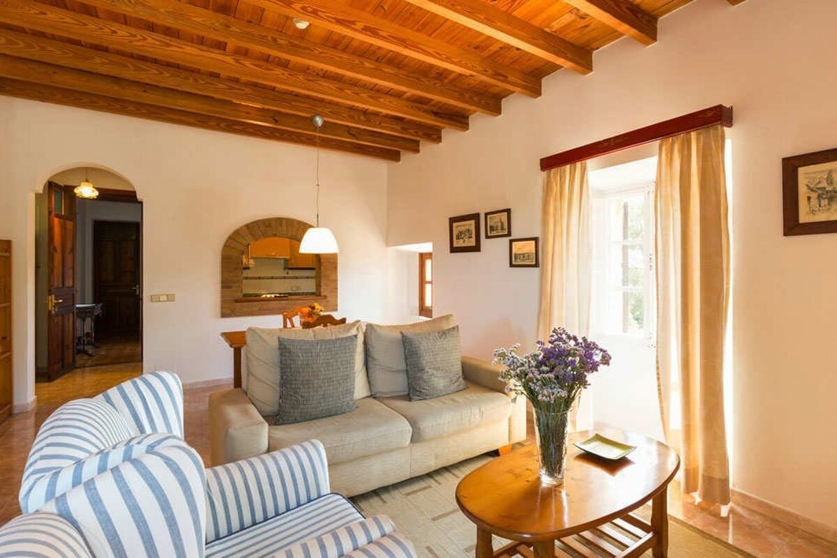 Cozy living room with a wooden beamed ceiling, two sofas, a coffee table with flowers, and an arched entryway to another room.