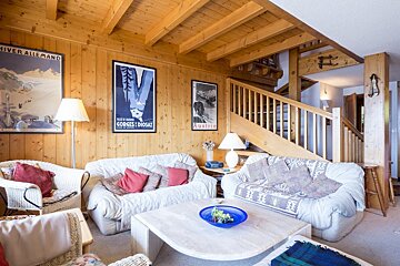 Warm, rustic chalet living room with wood-paneled walls, comfortable sofas, vintage ski posters, and a wooden staircase.
