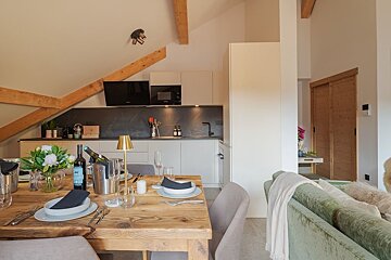 A modern open-plan living area featuring a rustic wooden dining table set for guests, a sleek kitchen, exposed ceiling beams, and a cozy green sofa.