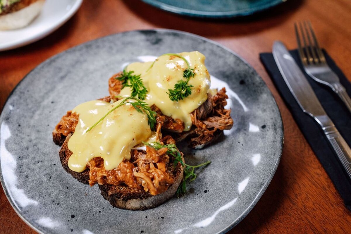 Two pulled pork Eggs Benedict with creamy hollandaise sauce and fresh herbs, served on a speckled grey plate with cutlery.
