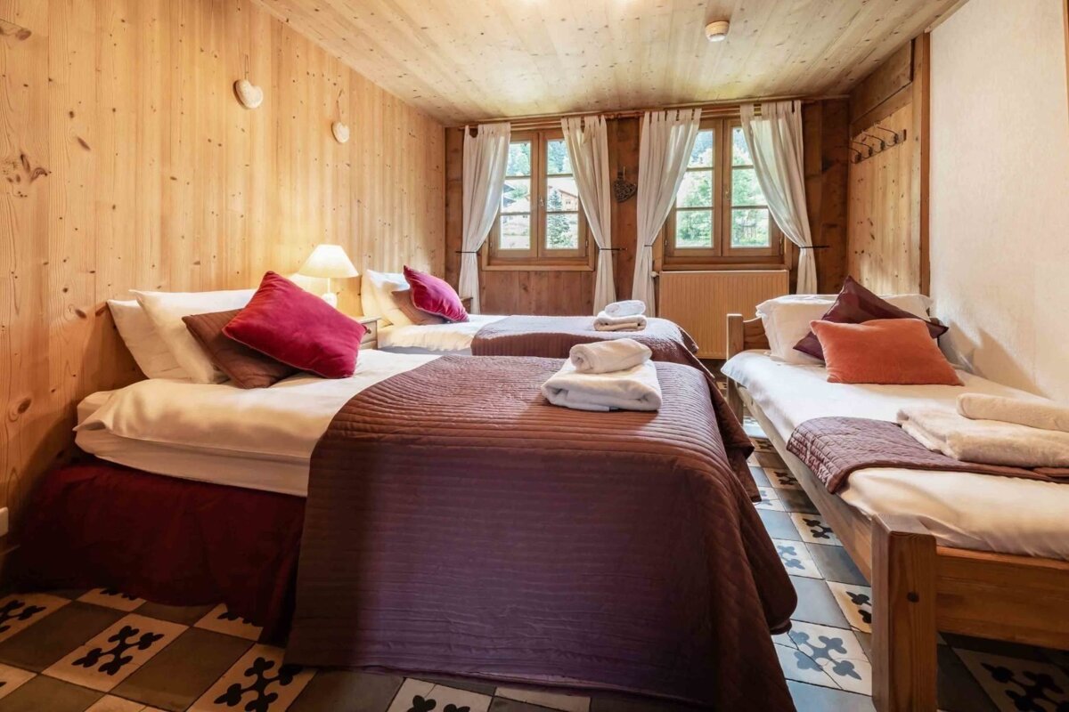 A rustic wooden bedroom features three beds with colorful pillows and white curtains. A patterned tiled floor adds character to the cozy space.