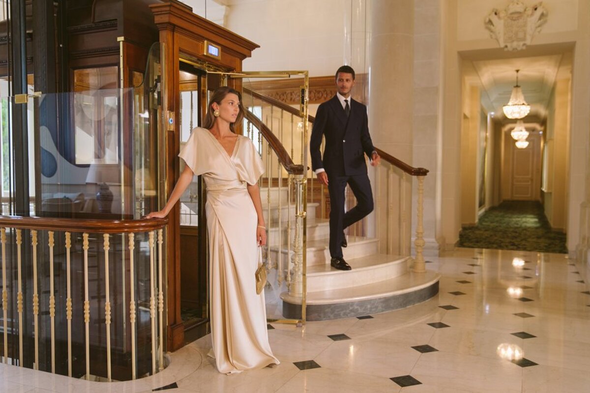 A woman in a white dress leans against a railing next to a man in a suit
