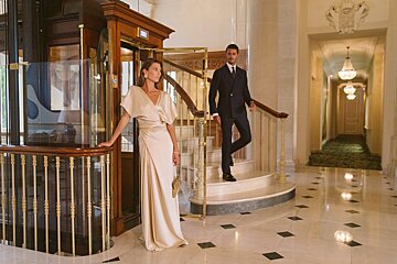 A woman in a white dress leans against a railing next to a man in a suit