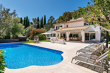 Sunny Mediterranean villa with a sparkling blue pool, extensive patio with loungers and outdoor furniture, set amidst green trees and distant hills.