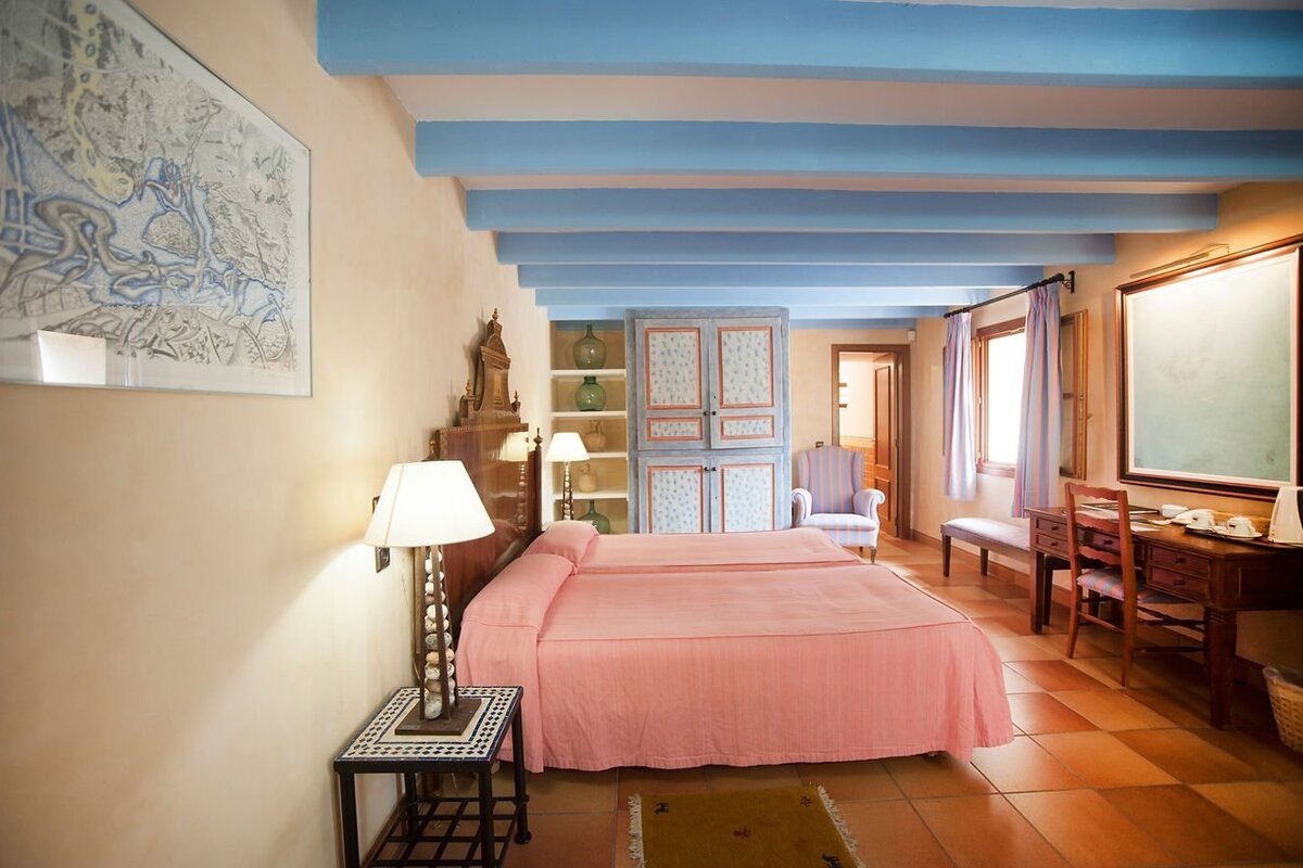 A cozy bedroom with two pink beds, blue ceiling beams, terracotta floor, and antique furniture, creating a rustic and inviting atmosphere.
