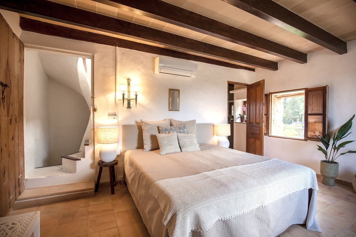 A rustic bedroom with exposed wooden beams, a comfy bed, tiled floor, an open window with shutters, and a unique spiral staircase.