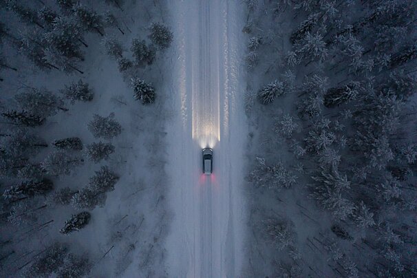 Aerial view of a car driving on a snow-covered road through a winter forest, its headlights illuminating the path ahead.