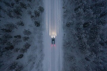 Aerial view of a car driving on a snow-covered road through a winter forest, its headlights illuminating the path ahead.