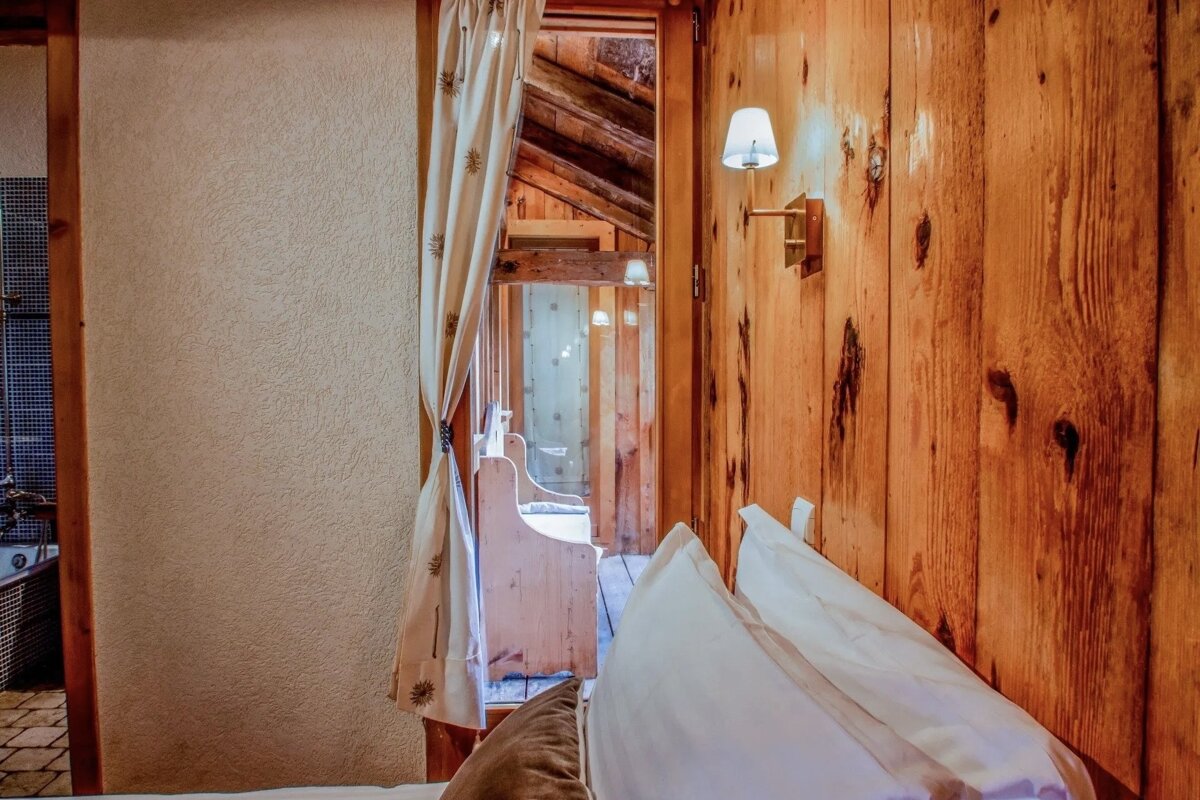 A cozy rustic bedroom view with warm wooden walls, a curtained doorway leading to a wooden hall, a glimpse of a bathroom, and pillows in the foreground.