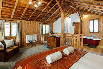 A charming, all-wood room with a cozy bed, sofa, and a striking red clawfoot tub. Vaulted wooden ceiling adds to the rustic appeal.
