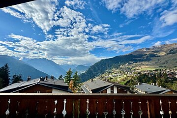 Panoramic view from a wooden balcony over a mountain village nestled in a valley, under a bright blue sky with scattered clouds.