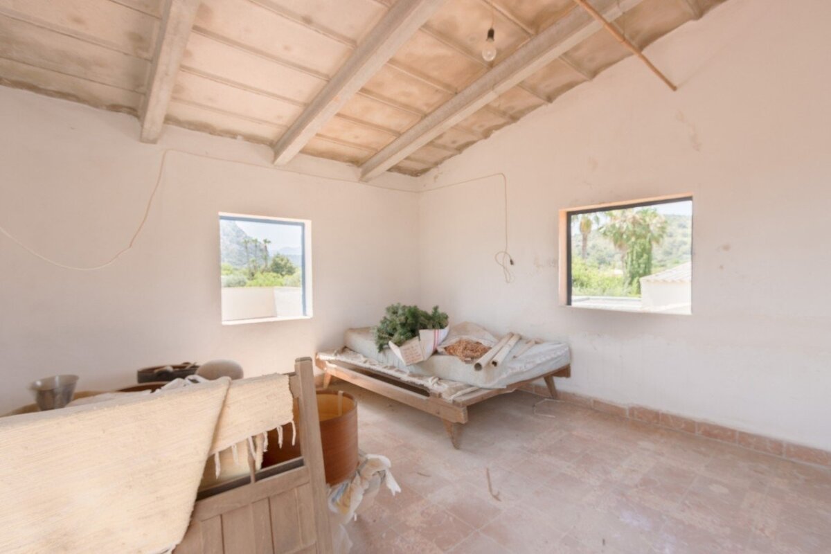 A rustic room with white walls, exposed ceiling, two windows revealing a natural landscape, a simple wooden bed, and tiled floor.