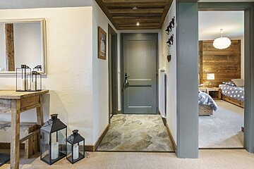 A rustic hallway with wooden ceiling beams and a stone floor leads to a grey door. An open doorway reveals a bedroom with wooden walls and two beds.