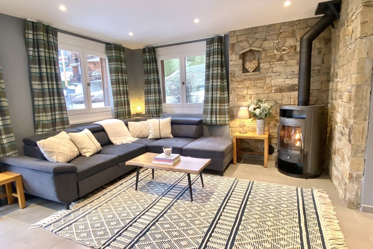 A cozy living room features an L-shaped sofa, plaid curtains, a stone accent wall with a wood-burning stove, a geometric rug, and a wooden coffee table.