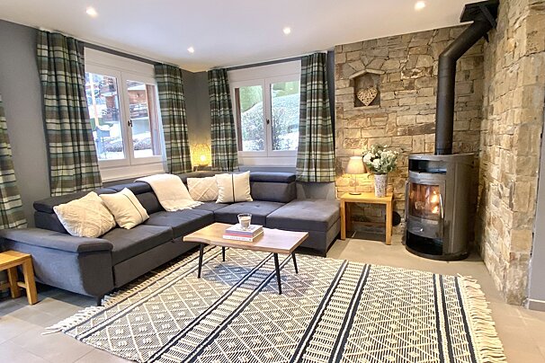 A cozy living room features an L-shaped sofa, plaid curtains, a stone accent wall with a wood-burning stove, a geometric rug, and a wooden coffee table.