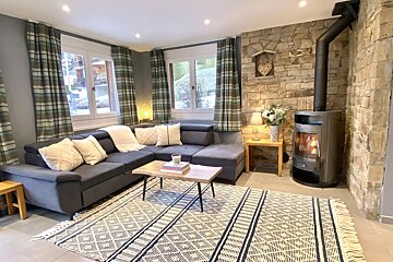 A cozy living room features an L-shaped sofa, plaid curtains, a stone accent wall with a wood-burning stove, a geometric rug, and a wooden coffee table.