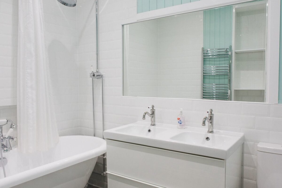 A modern, white tiled bathroom featuring a tub with shower, double vanity, and a large mirror reflecting a blue wall and heated towel rack.