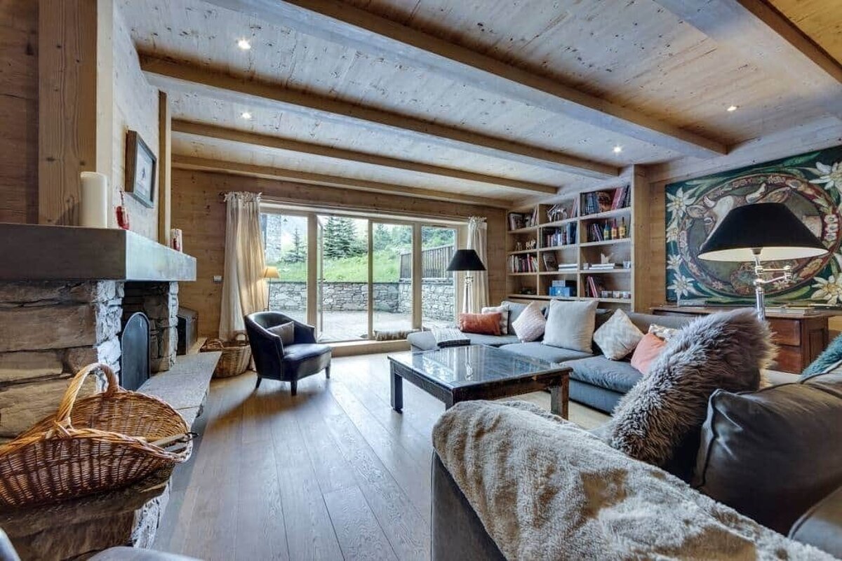 Cozy, rustic living room with wood-paneled walls/ceiling, stone fireplace, comfy sofas, bookshelf, and glass doors opening to a green outdoor view.
