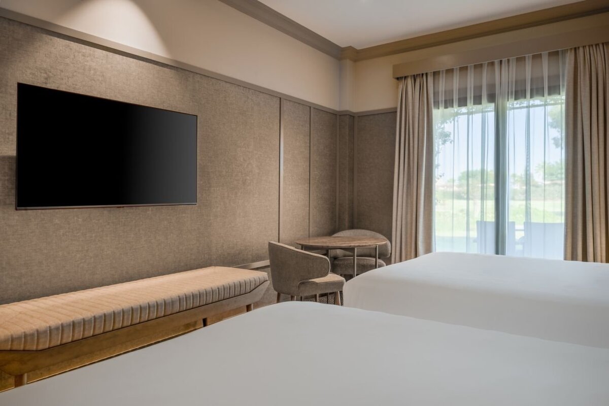 A modern hotel room with two beds, a large TV, a bench, a small table with chairs, and a window overlooking a green landscape.