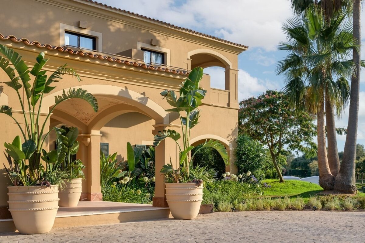 A Mediterranean-style building with arches, large potted plants, and palm trees under a blue sky, surrounded by lush landscaping.
