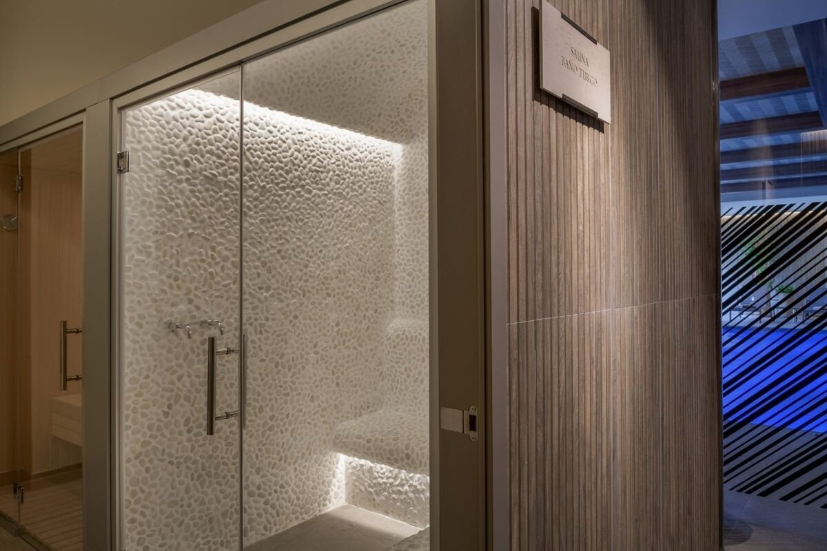 Modern sauna/steam room with pebble-textured walls, glass doors, ambient lighting, and a wooden wall, beside a patterned glass revealing a blue pool.