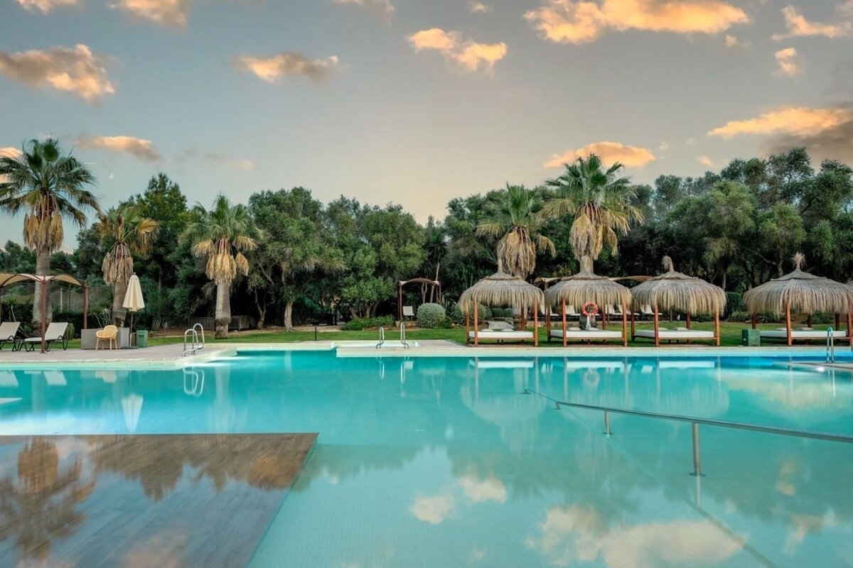 A serene resort pool with clear blue water, surrounded by palm trees, lush greenery, and thatched sunbeds under a cloudy sky.
