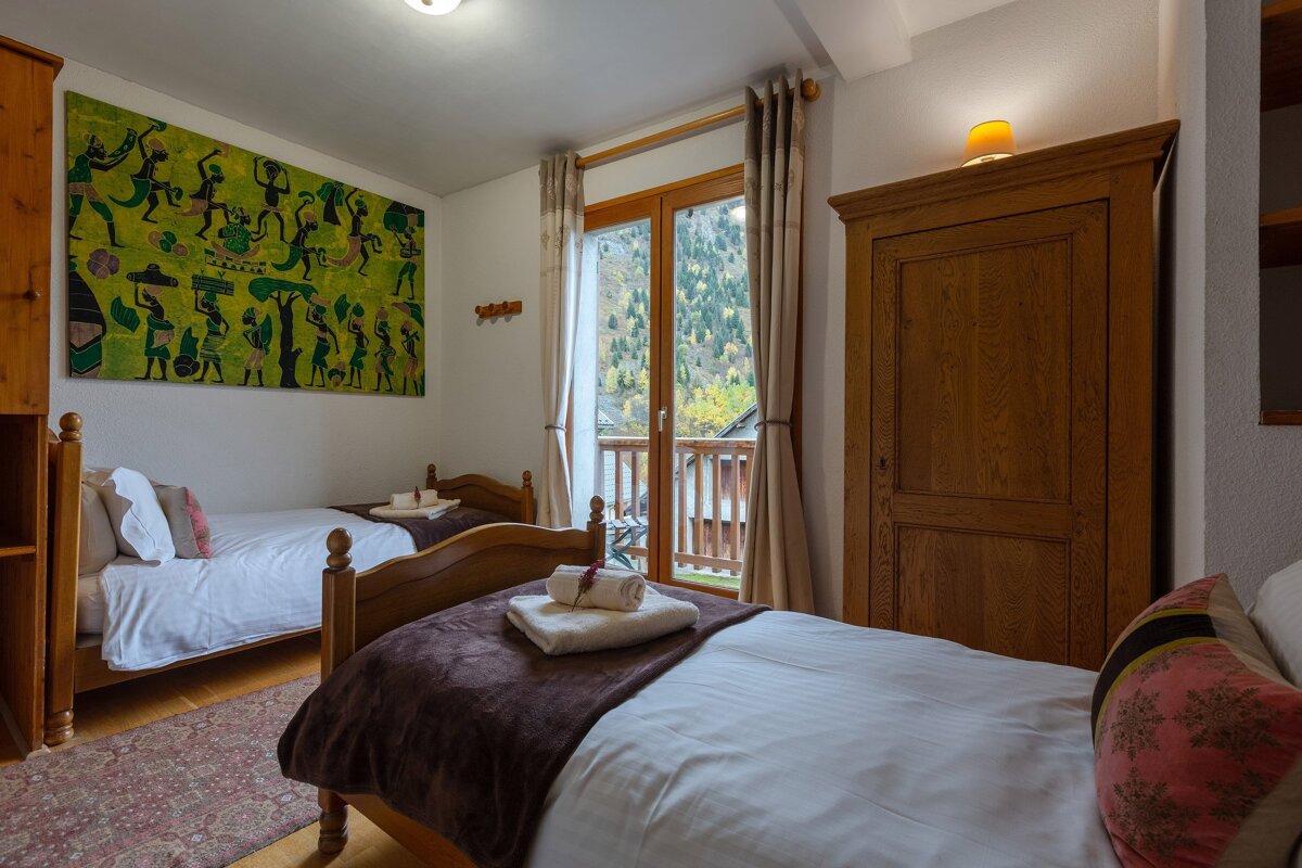 A cozy alpine twin bedroom with wooden beds, vibrant green artwork, and a balcony offering a picturesque view of a lush, forested mountain.