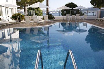 A resort swimming pool with a ladder and reflections of umbrellas, surrounded by lounge chairs, overlooking a bay with boats.