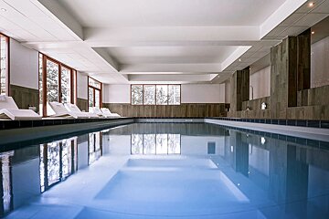 A bright indoor swimming pool with still blue water. White lounge chairs line the side, and large windows offer views of a snowy, forested landscape.