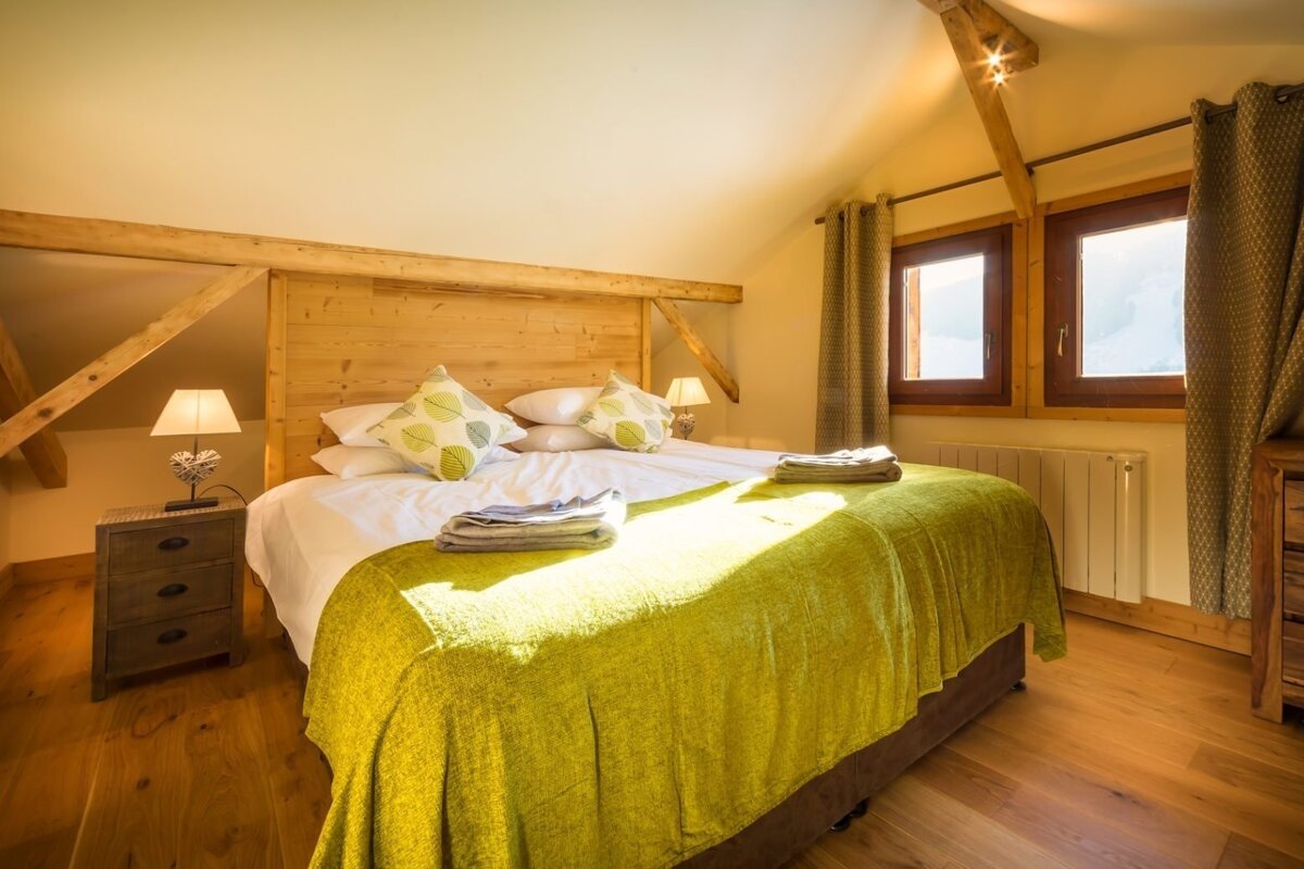 A cozy, sunlit bedroom features a large bed with a green blanket, wooden headboard, and exposed beams. Light streams through windows onto the hardwood floor.