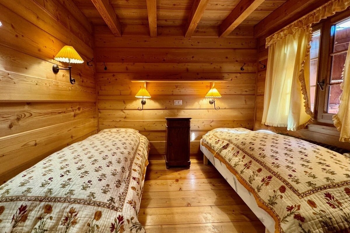Cozy, all-wood cabin bedroom with two single beds, floral quilts, warm wall lamps, and a window with yellow curtains.