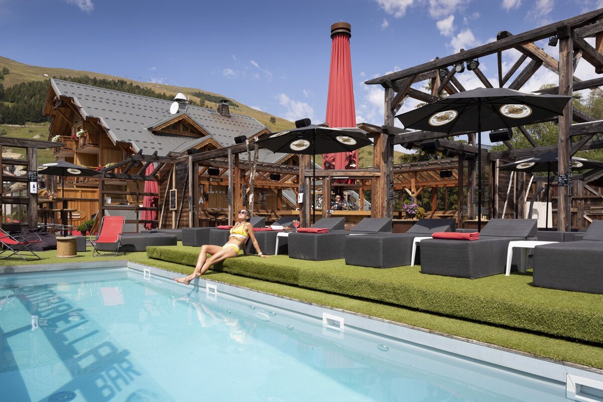 A woman sits on the edge of a swimming pool in front of a hotel bar