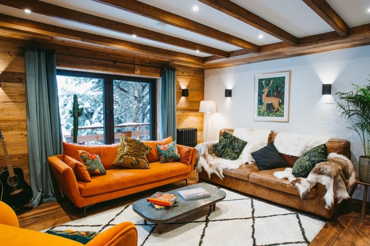 A cozy, modern chalet living room featuring wood paneling, an orange sofa, a brown leather sofa, and a large window with a snowy view.