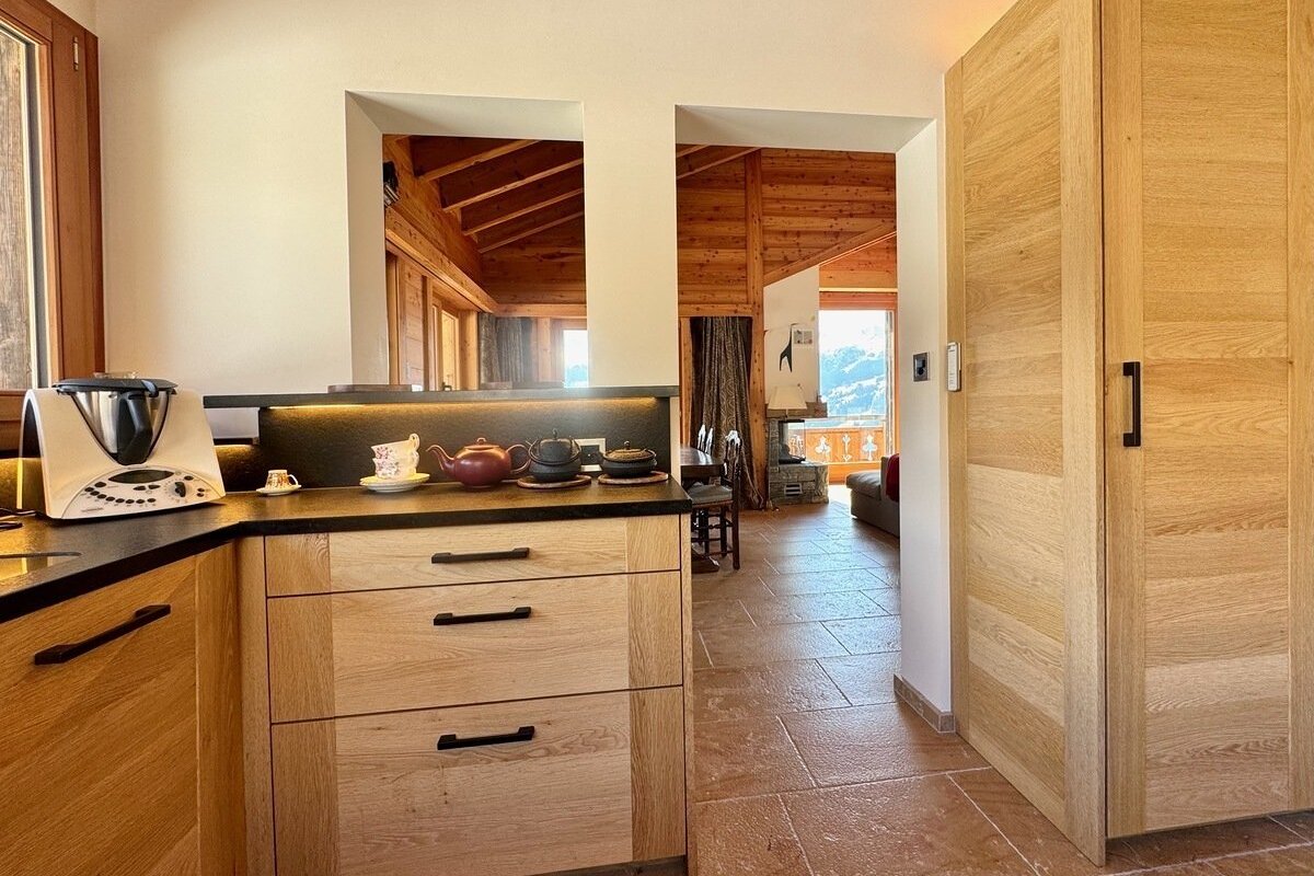 Modern wooden kitchen with a Thermomix and teapots, looking into a rustic living area with a fireplace and mountain views.