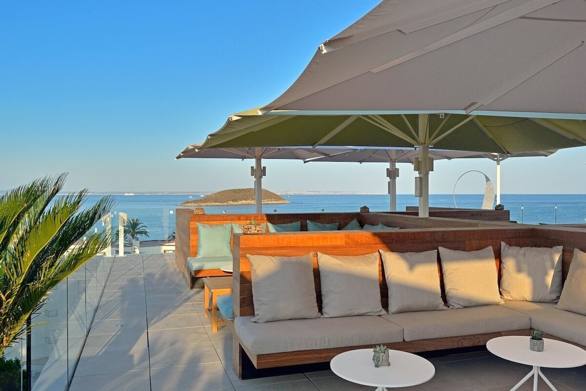 A chic rooftop terrace with lounge seating and large umbrellas offering panoramic views of a tranquil blue sea, distant islands, and a clear sky.